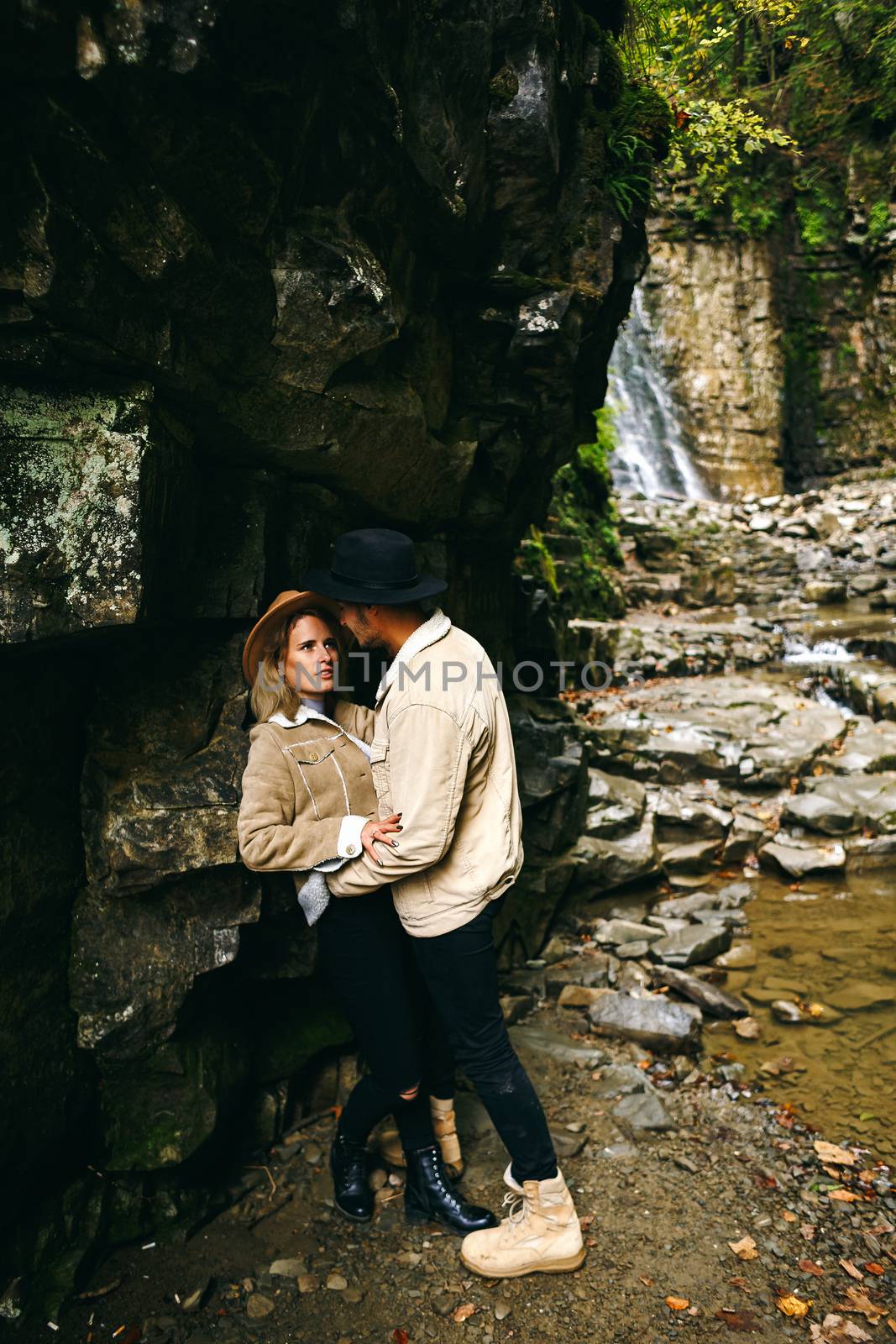 Young and beautiful couple at the mountain waterfall - Happy tourists visiting mountains. Lovestory. Tourists in hats. Military fashion. by Denys_N