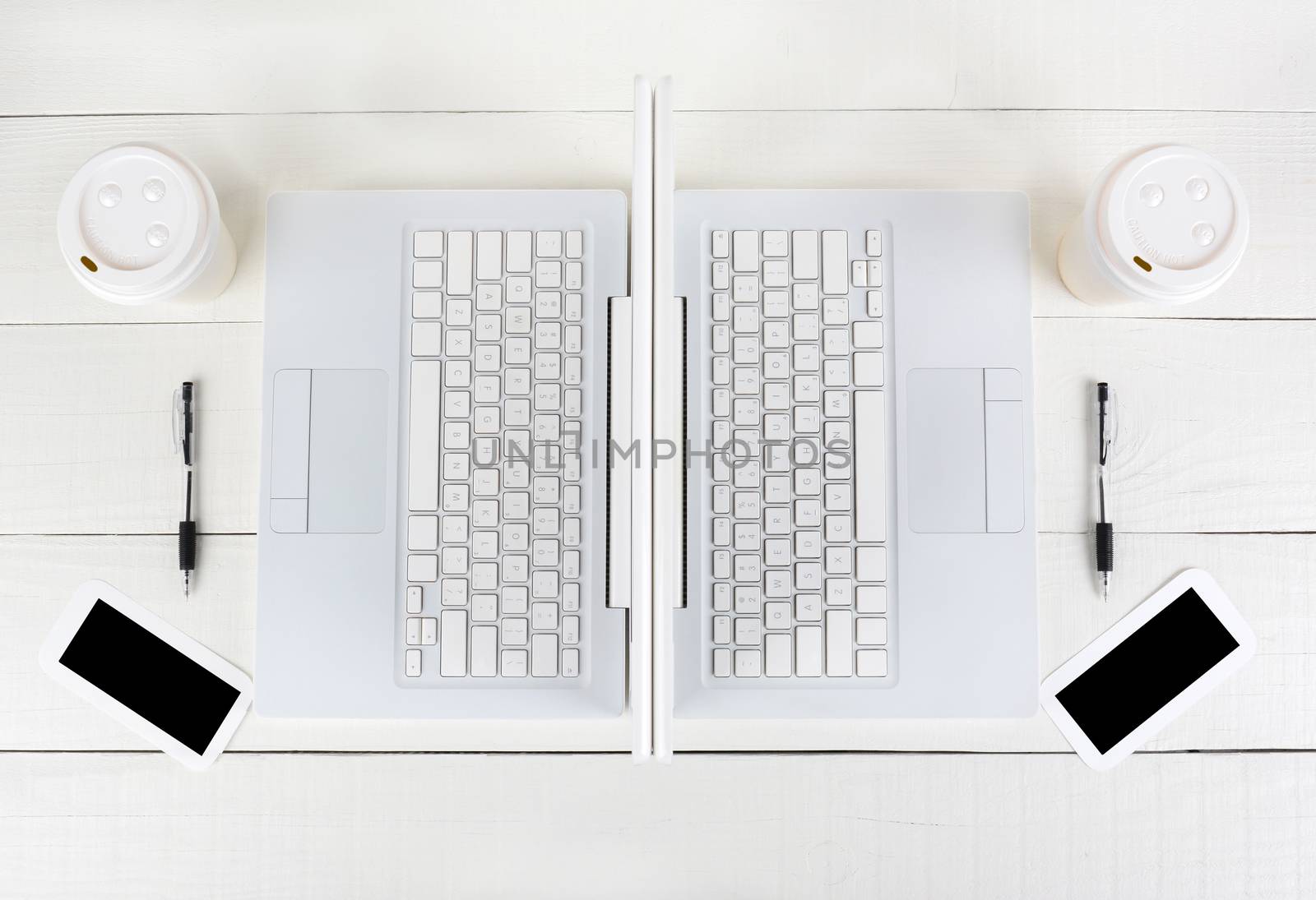 High angle shot of two laptop work stations back-to-back with almost identical layouts. Horizontal format on a white wood table.