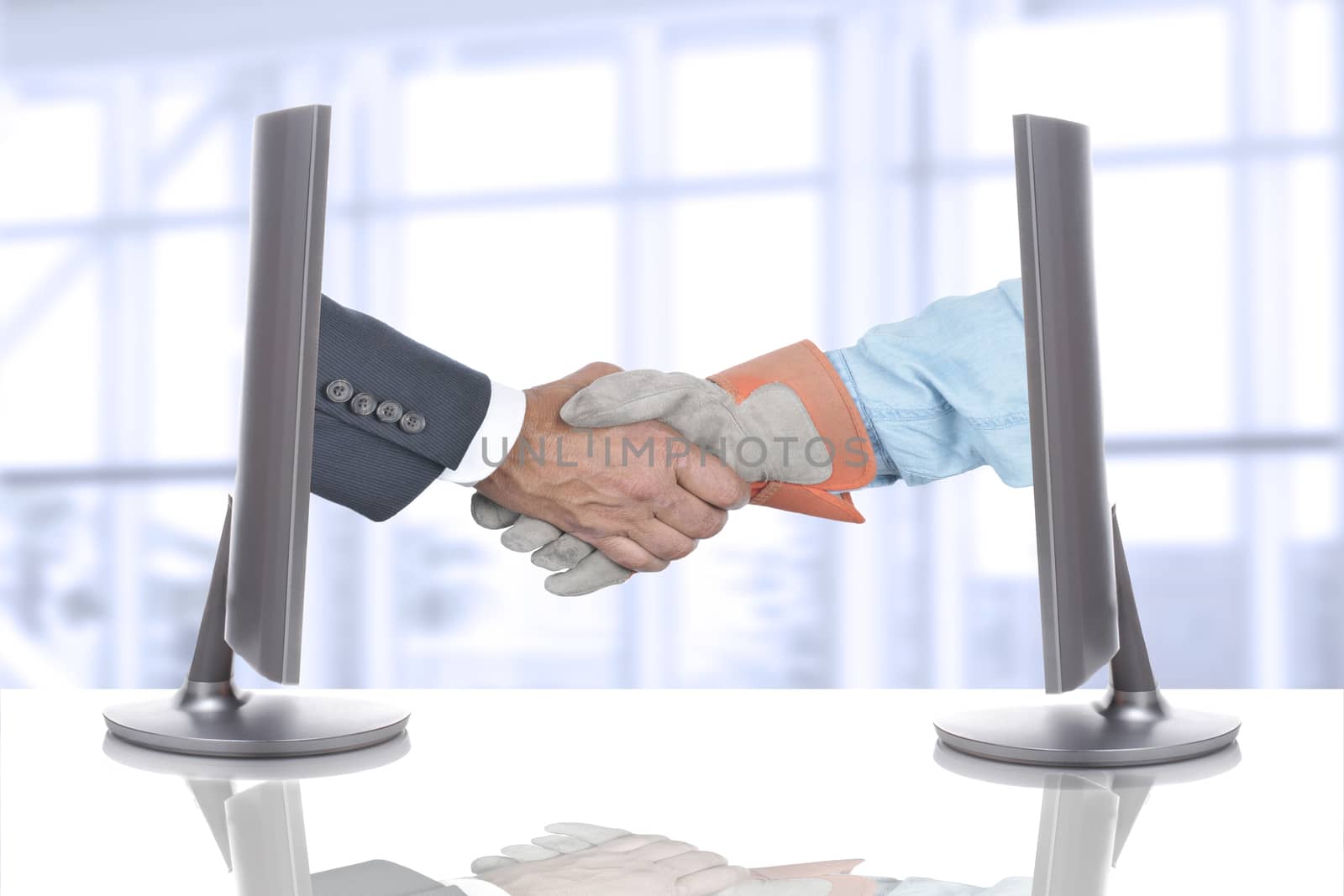 Handshake Over Desk in Business Office. Both hands are coming out of computer screens. 
