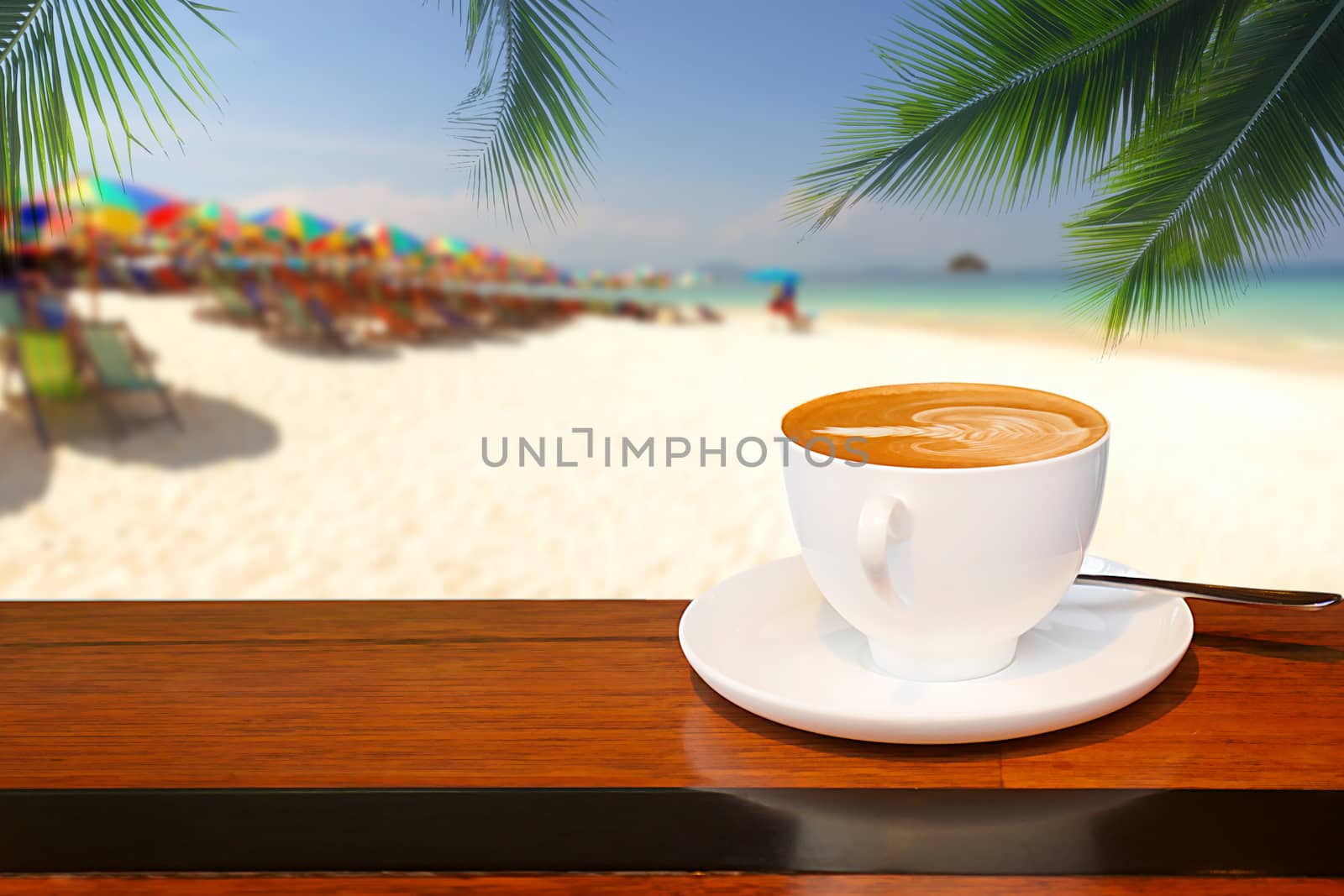A cup of coffee resting on a wooden table with a coconut leaf background and blurred beach by chiawth