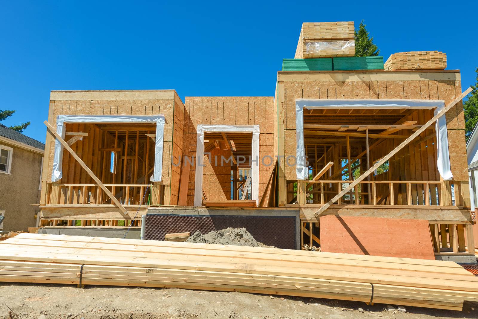 A single family home under construction. The house has been framed and covered in plywood. Stacks of board timber in front and stack of 2x4 boards on the top.