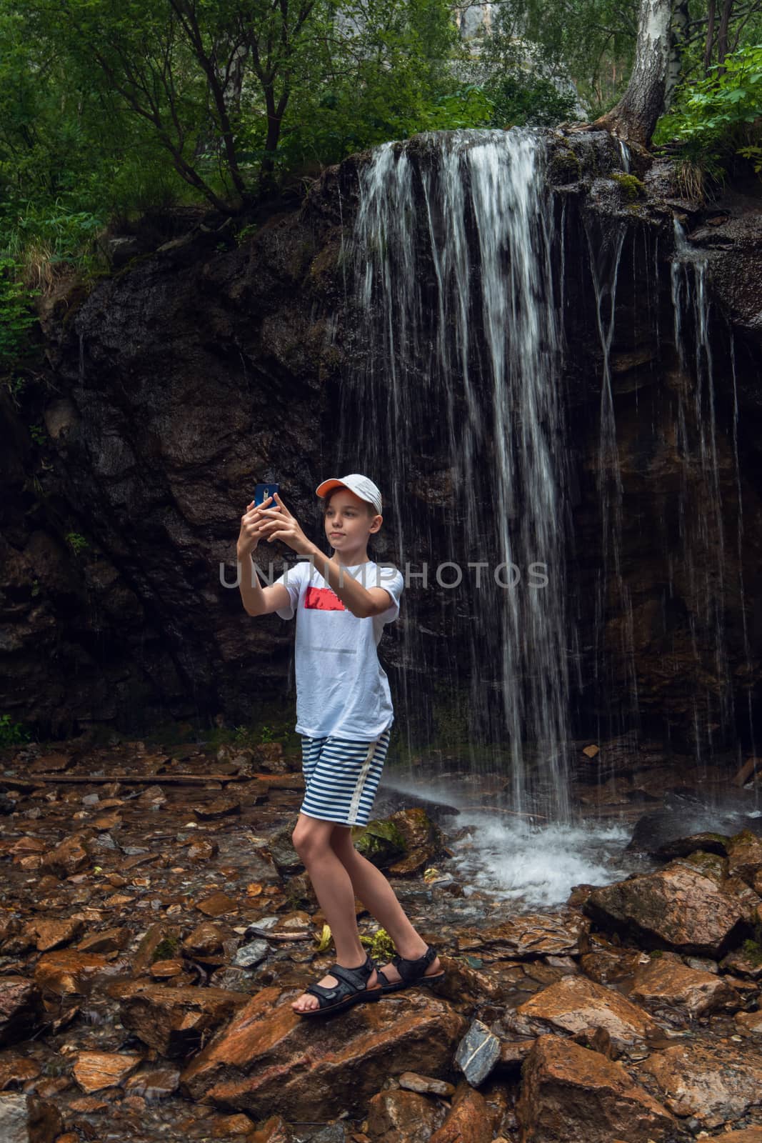 Stunning view of a boy tourist taking selfie photos with a smartphone at the waterfalls background in the Altai mountains.