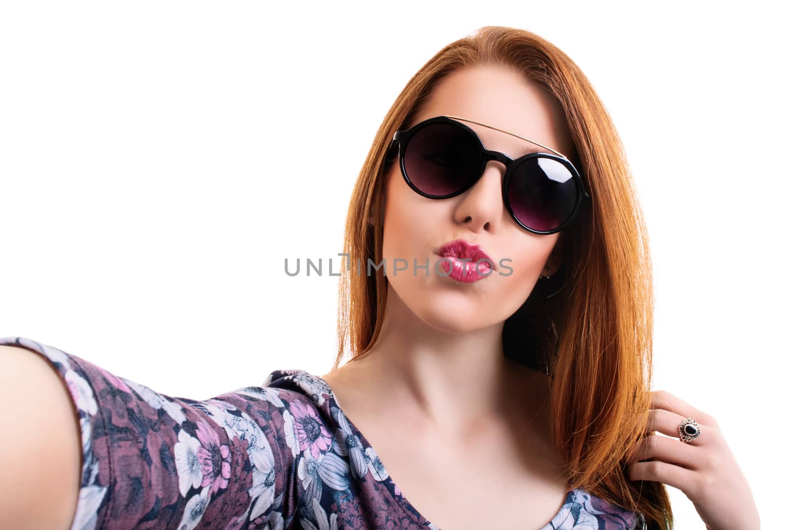 Young beautiful girl with sunglasses taking a selfie while blowing a kiss, looking at the camera POV, isolated on white background. Attractive redhead girl with long hair taking a selfie. Lifestyle, happiness, and social concept.