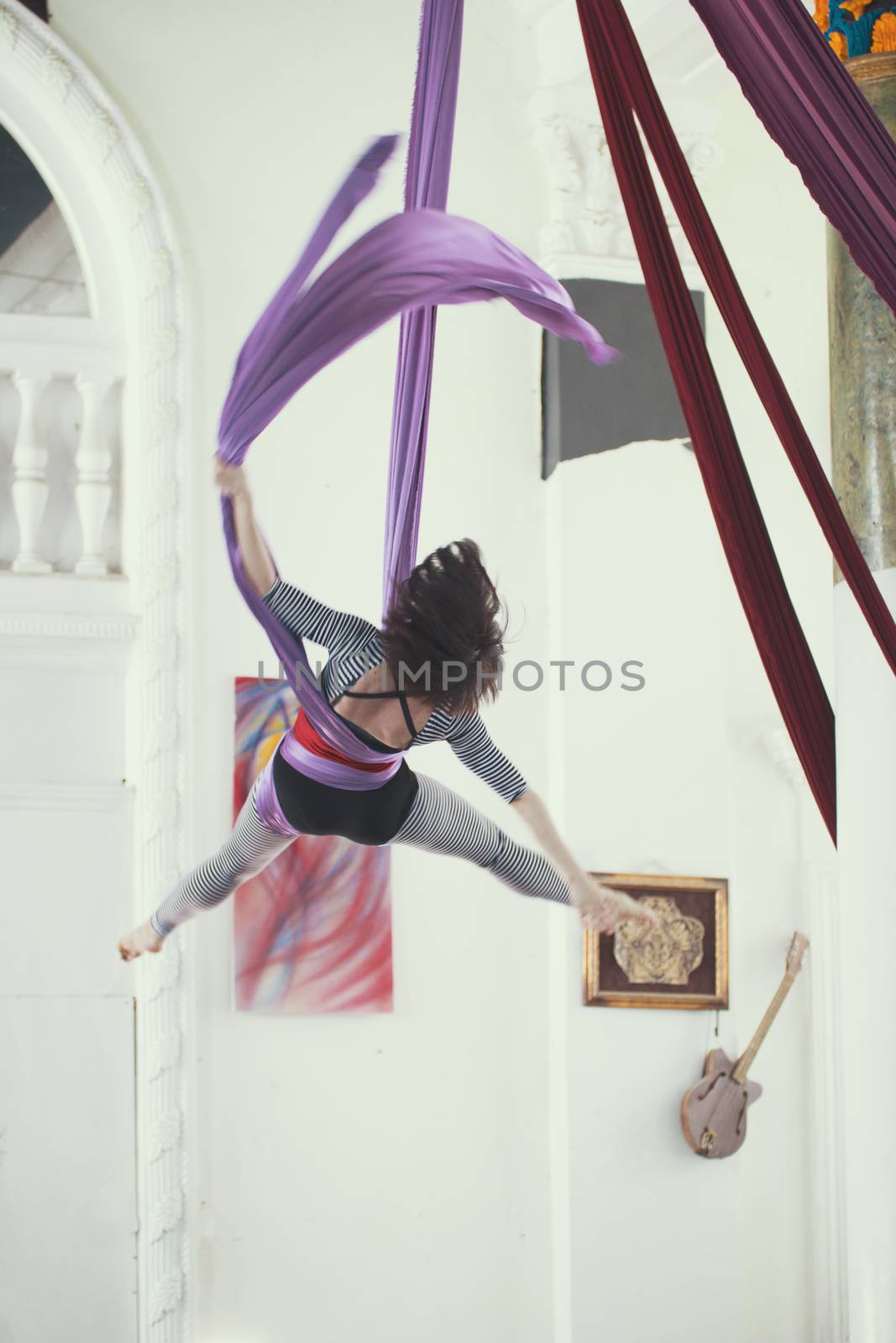 Flexible young woman flying on the aerial silk is a bright studio, telephoto shot