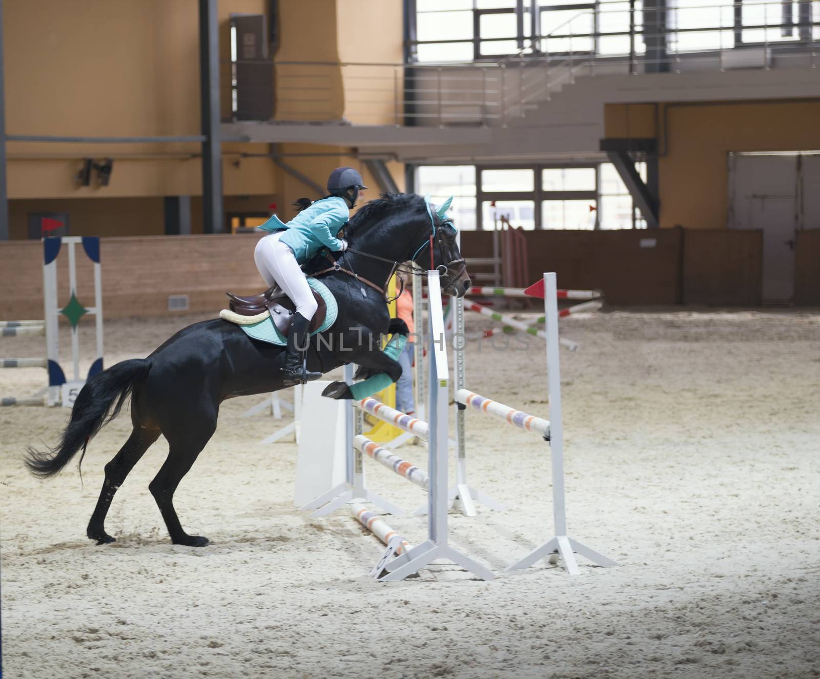 Young woman on the black stallion jumping over hurdle at show jumping competition by Studia72