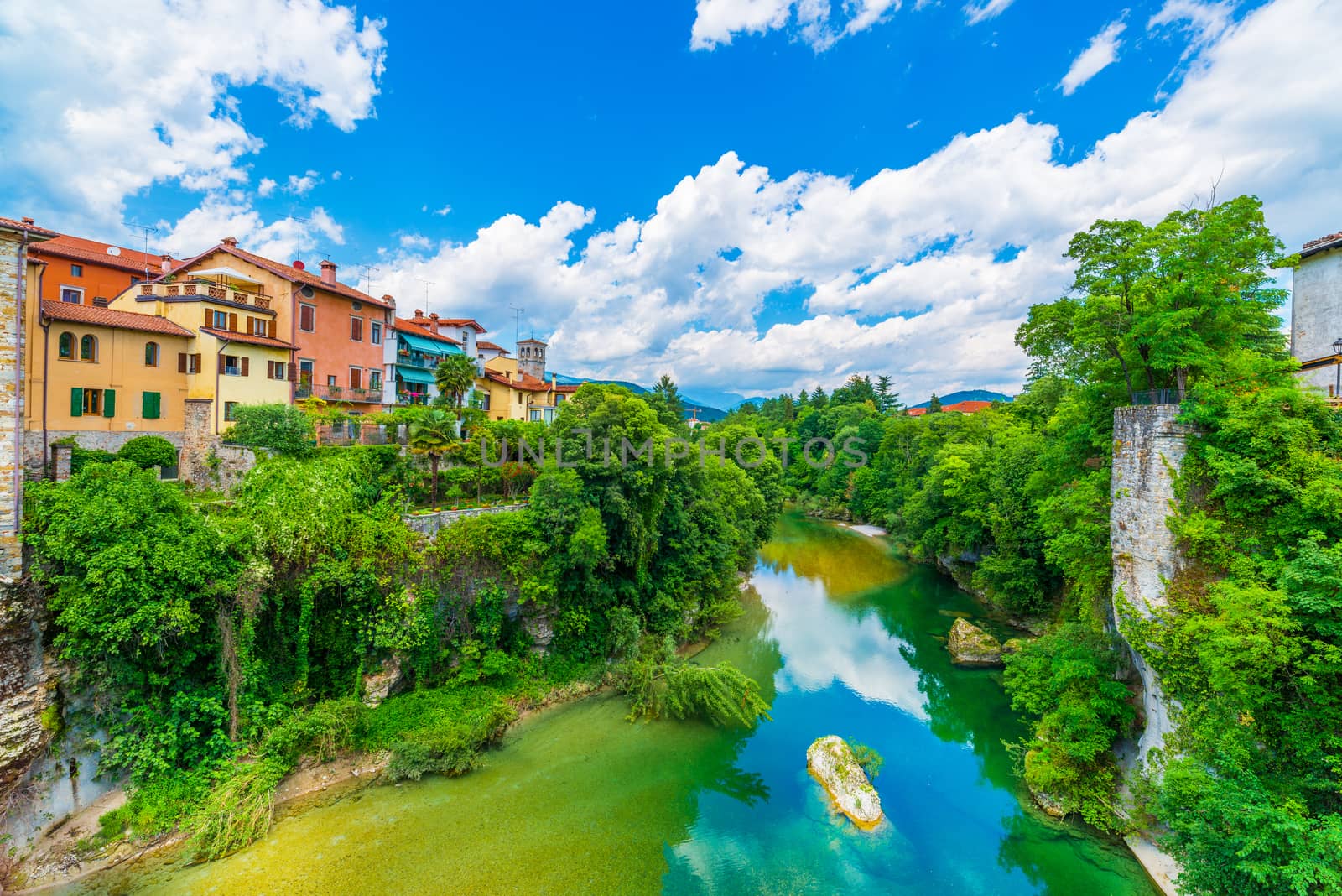 Old Italian city of Cividale del Friuli. Medieval town in Northern Italy. Cityscape with colored residential houses, river, cliffs with green trees and blue sky with clouds.