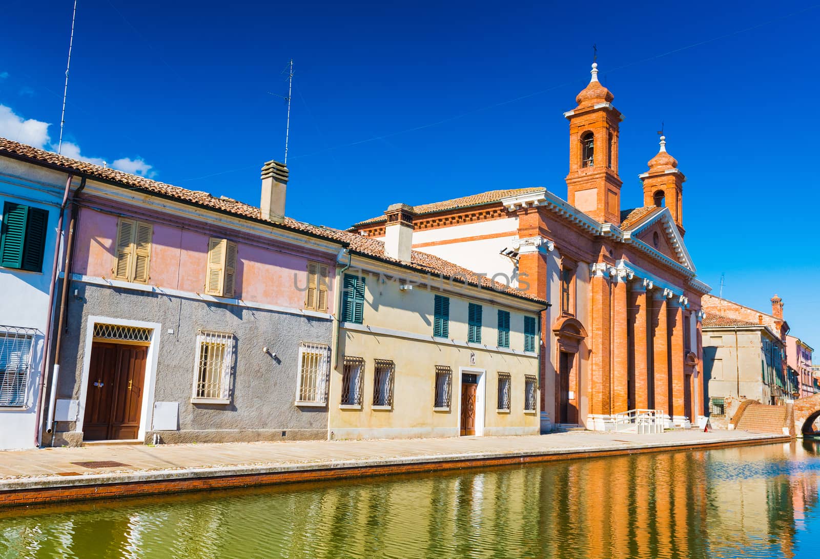 View of the street with canal and colored houses in a small Italian town of Comacchio (Little Venice). San Camillo Hospital (museum of the Roman Ship) against the clear blue sky, Emilia-Romagna, Italy