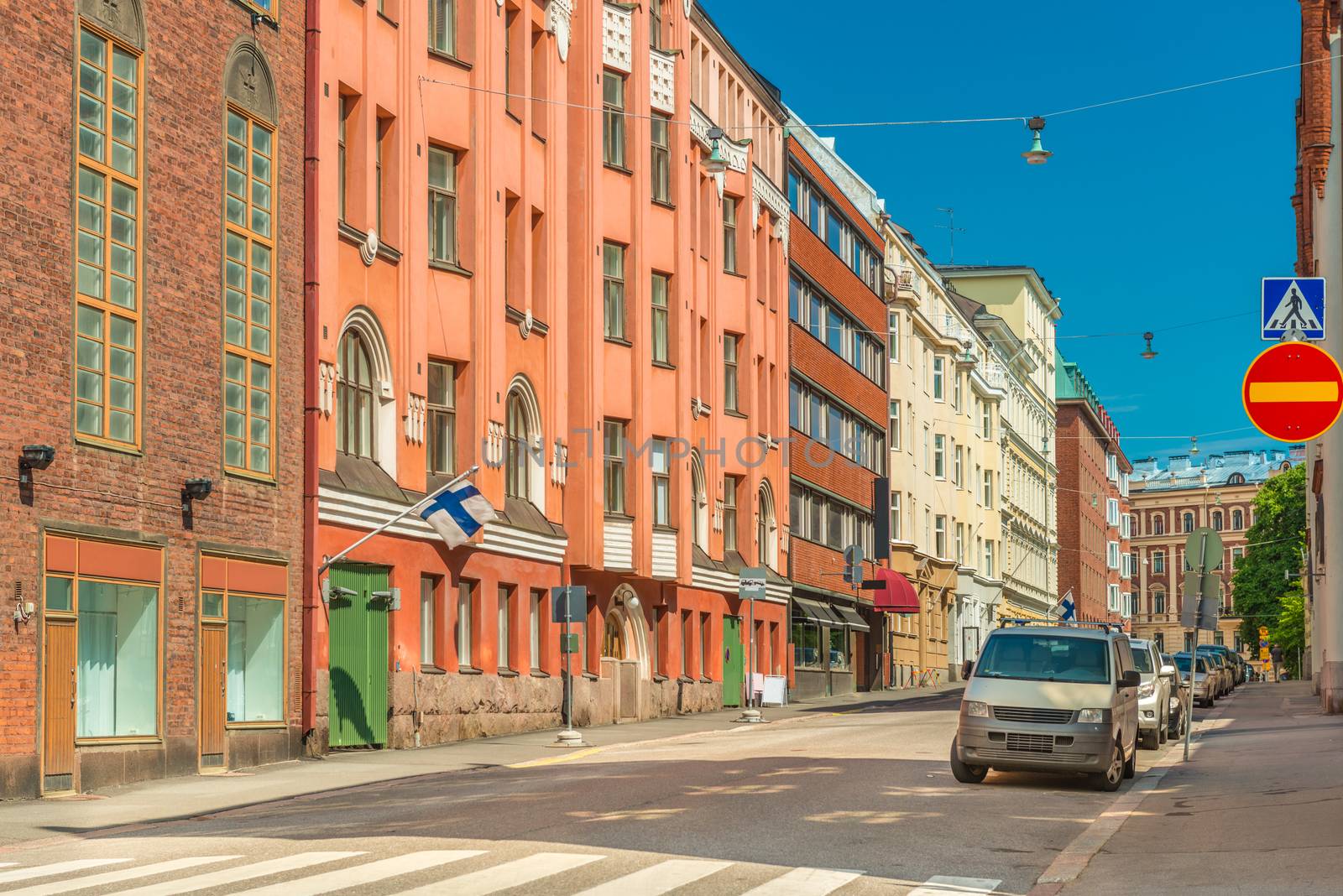 View of an empty street in Helsinki, Finland. Colored historical buildings with Finnish flags on the facades, parked cars and clear blue sky