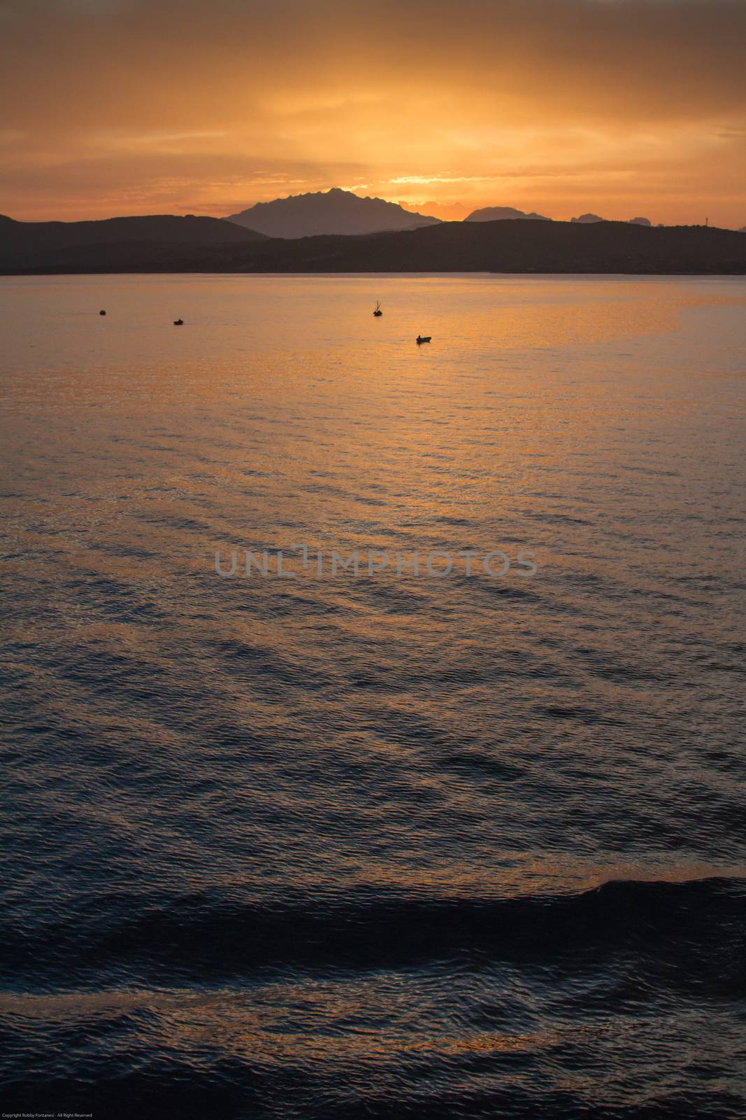 Sunrise on the Sardinian sea coast with intense orange color seen from the sea with five fishing boats on flat water