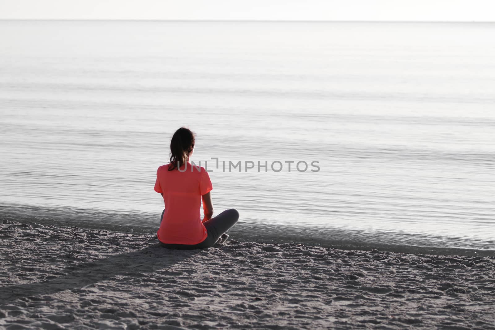 Young woman sitting on the shoreline of the sea at dawn in athletic outfit, looks towards the sea illuminated by the low sun and meditates