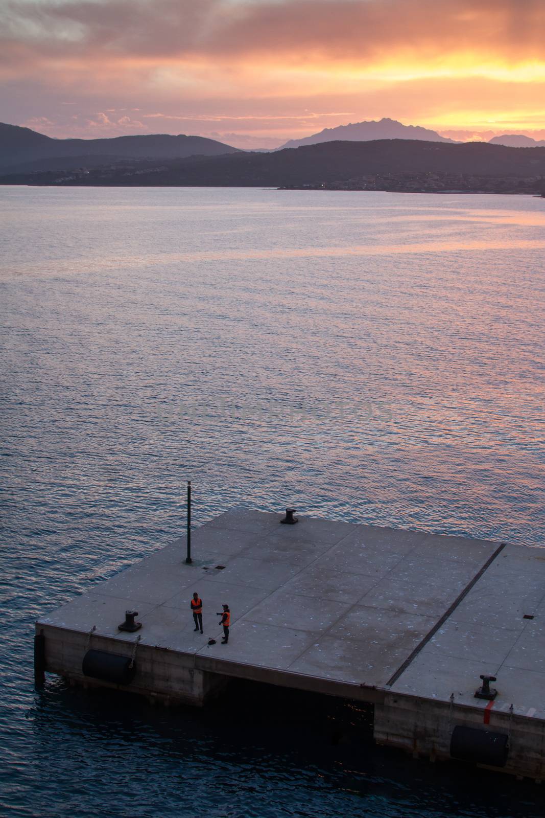 Sunrise on the Sardinian sea coast with intense orange color seen from the sea on the ferry that is about to dock and two workers of the port on the pier