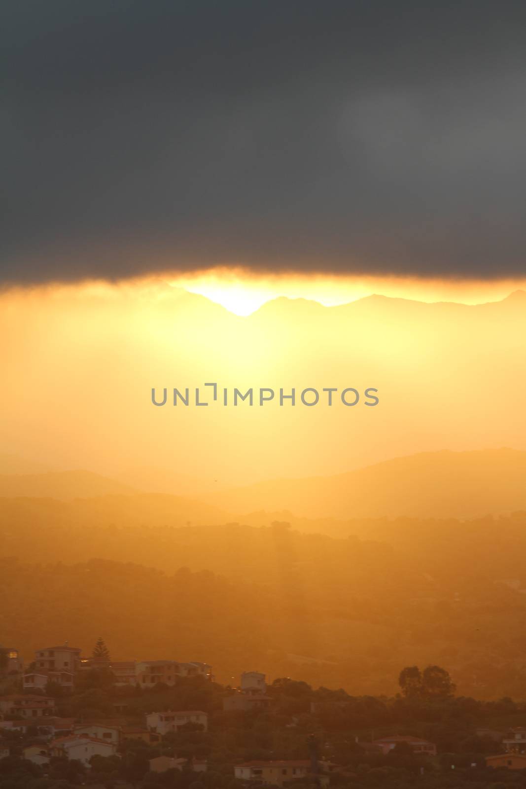 Breathtaking sunset after the storm. The orange sun's rays slip under a big black cloud illuminating the mountains and the hills with the houses of a small village by robbyfontanesi