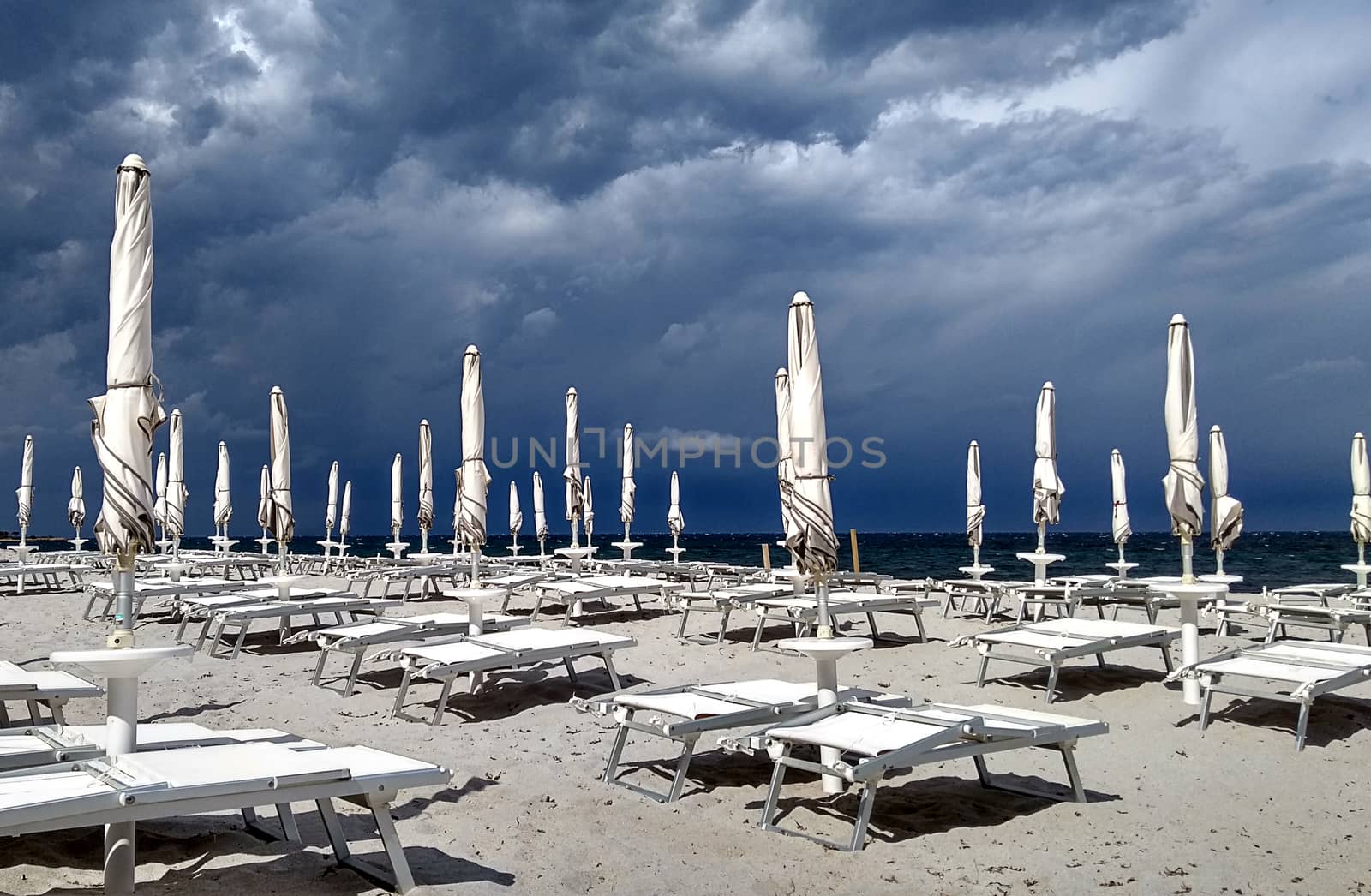 Dark and stormy sky, with gray clouds on the blue sea and a little beach with deck chairs and closed white umbrellas in the foreground by robbyfontanesi