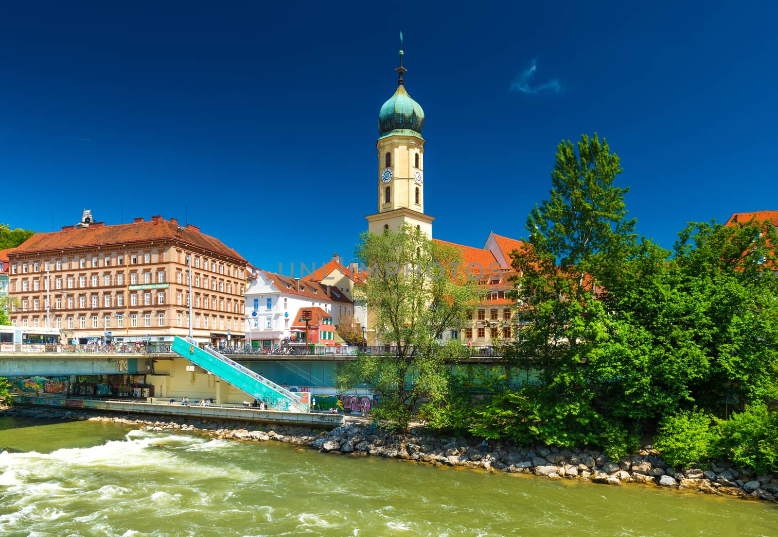 Graz - May 2017, Austria: The old city center on sunny day. Franciscan Church, old historical buildings with orange roofs against the blue sky and the flowing river Mur