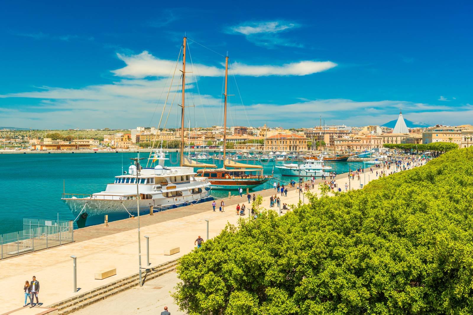 Syracuse - April 2019, Sicily, Italy: Cityscape of Syracuse viewed from Ortygia. View of an embankment with walking people, turquoise water, yachts and touristic ships