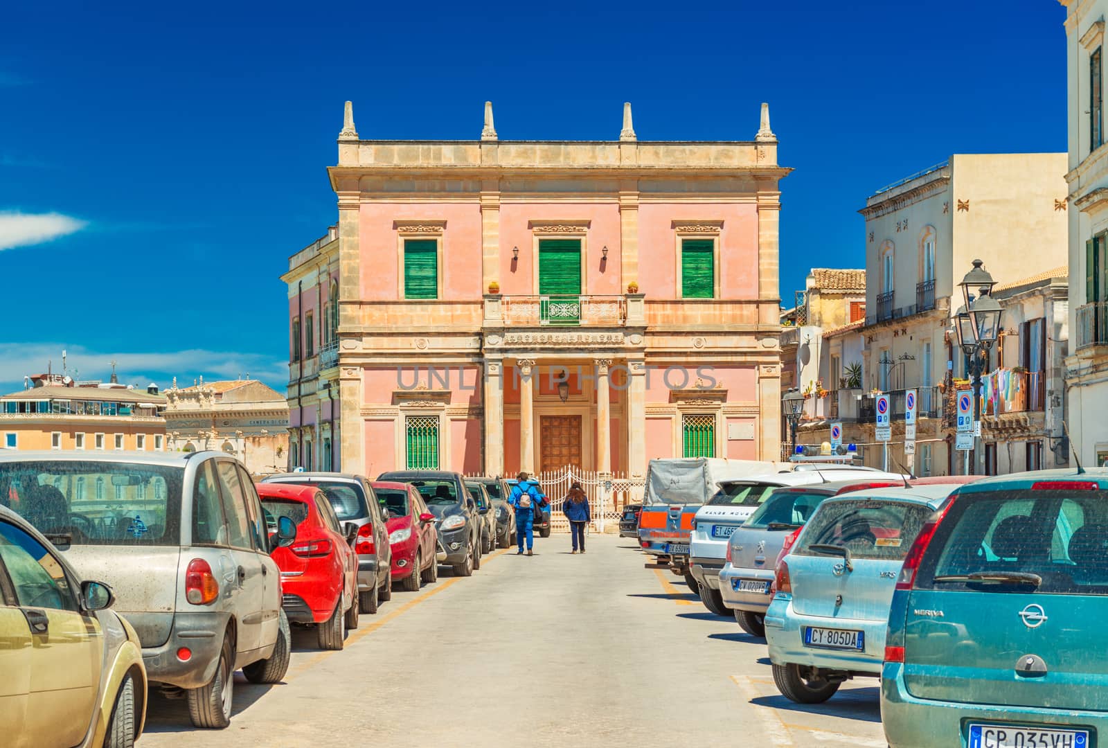 Syracuse - April 2019, Italy: View of an old historical building with a couple of tourist walking towards it. Cars parked along the street
