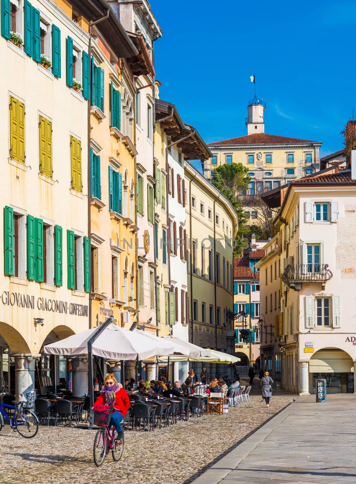 Udine - February 2017, Northern Italy: Woman in red jacket is cycling in the center of the city. The street of Udine with colored houses in traditional architectural style. People are sitting in cafe