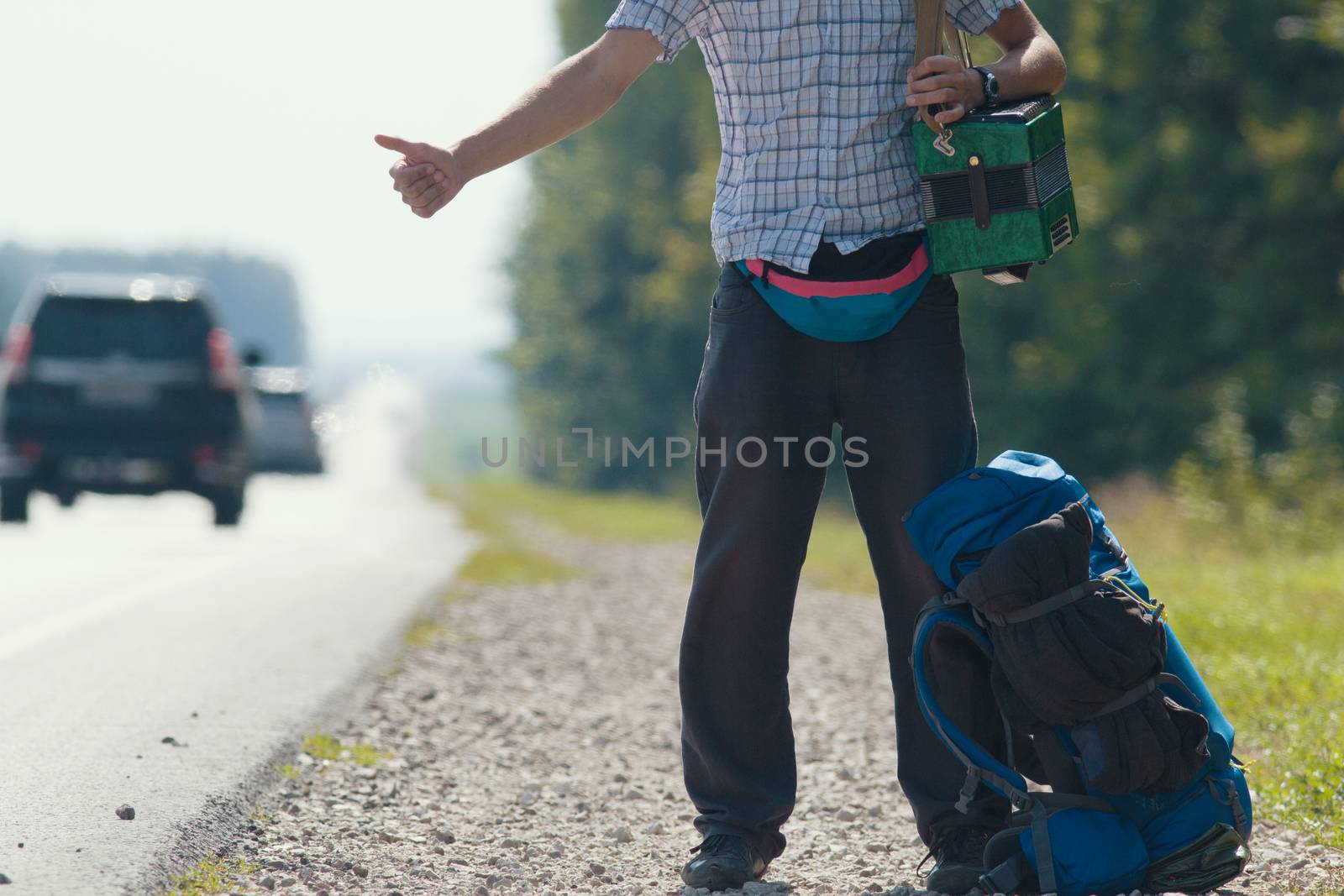 A man in a plaid shirt, dark pants, with an accordion on his shoulders standing by the road and honking, backpack lying at his feet by Studia72