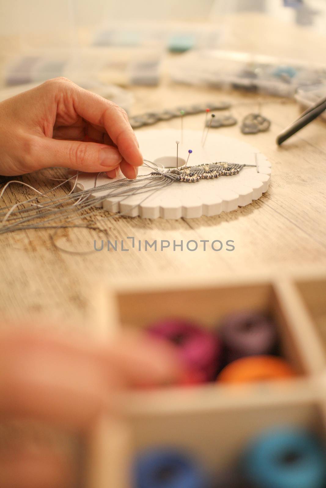Close up in selective focus of female hands making a macramé bracelet with kumihimo on a wooden table with tools, spools of thread, natural stones and colored beads by robbyfontanesi