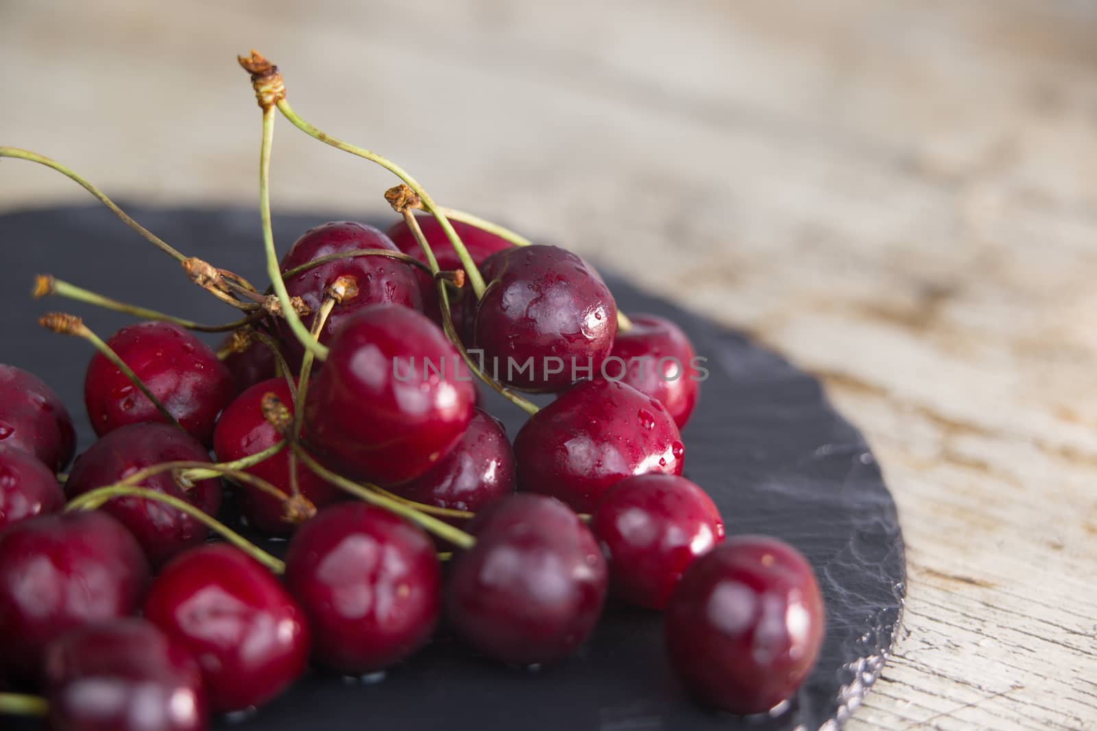 Fresh cherries on a black plate of wet slate on light wooden table in selective focus for copy space by robbyfontanesi