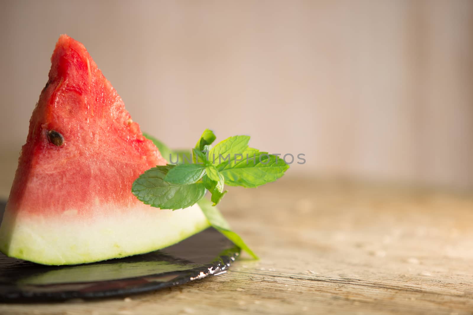 Triangular slice of watermelon standing on black plate of wet slate with sprig of fresh green mint in selective focus for copy space by robbyfontanesi