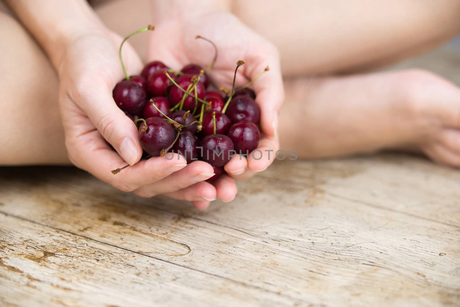 Woman sitting cross-legged on the light wooden table with her hands together holding a large handful of cherries by robbyfontanesi