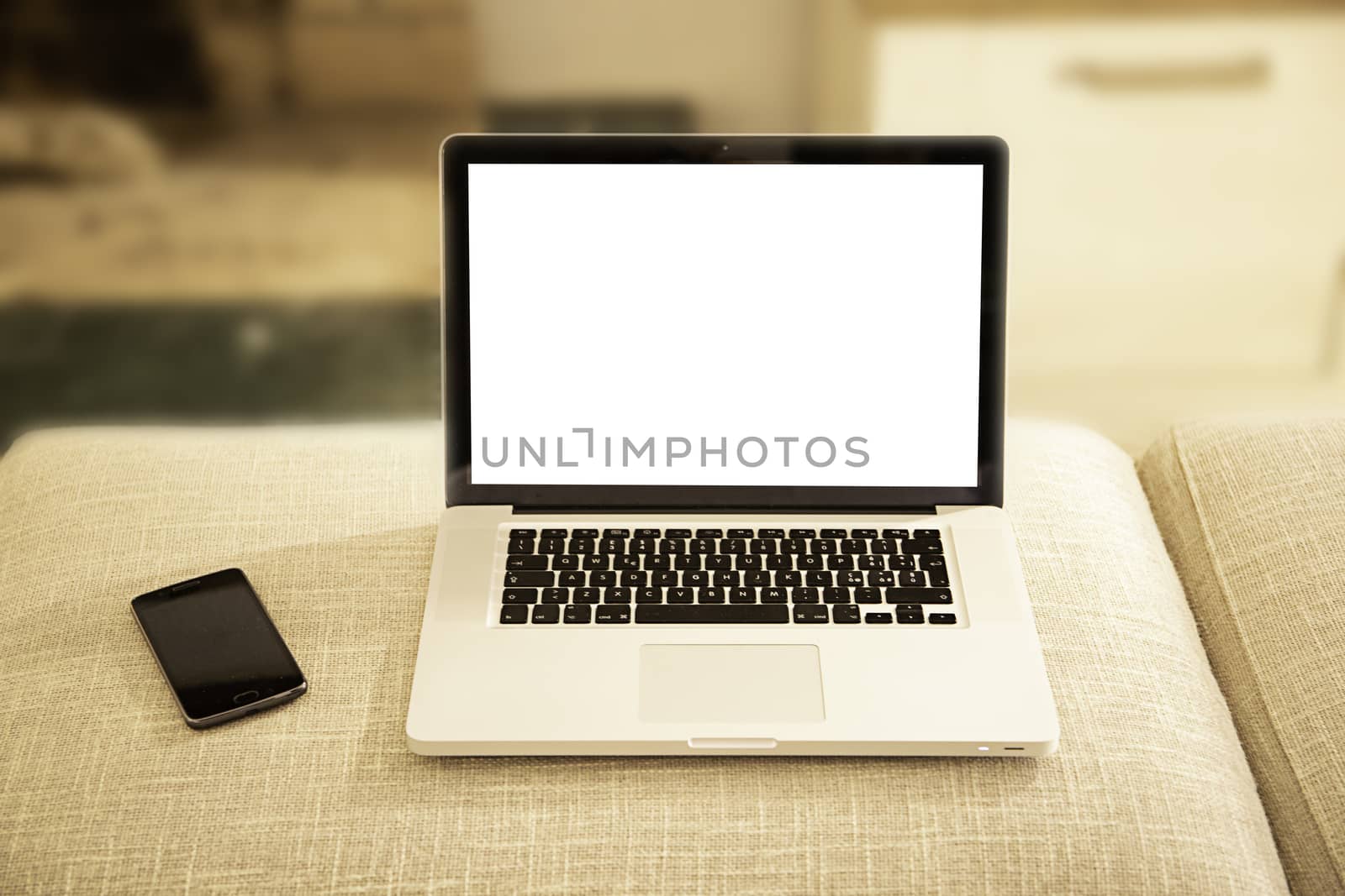 Open aluminum laptop with blank screen for copy space and smartphone on a sofa ottoman in selective focus with shaded lounge background