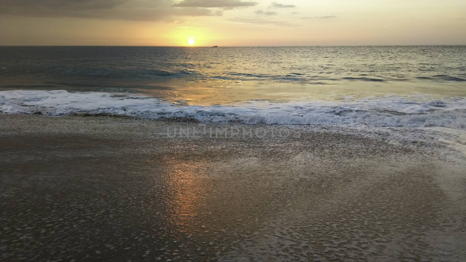 Sunset over the ocean from the shore of Nazaré beach, Playa do Norte, Portugal. The waves break on the shore reflecting the colors of the sky on the wet sand by robbyfontanesi