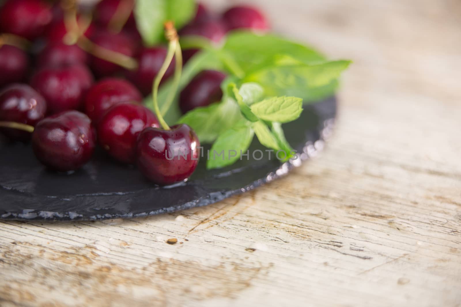 Fresh cherries on a black plate of wet slate with sprig of fresh green mint on light wooden table in selective focus for copy space by robbyfontanesi