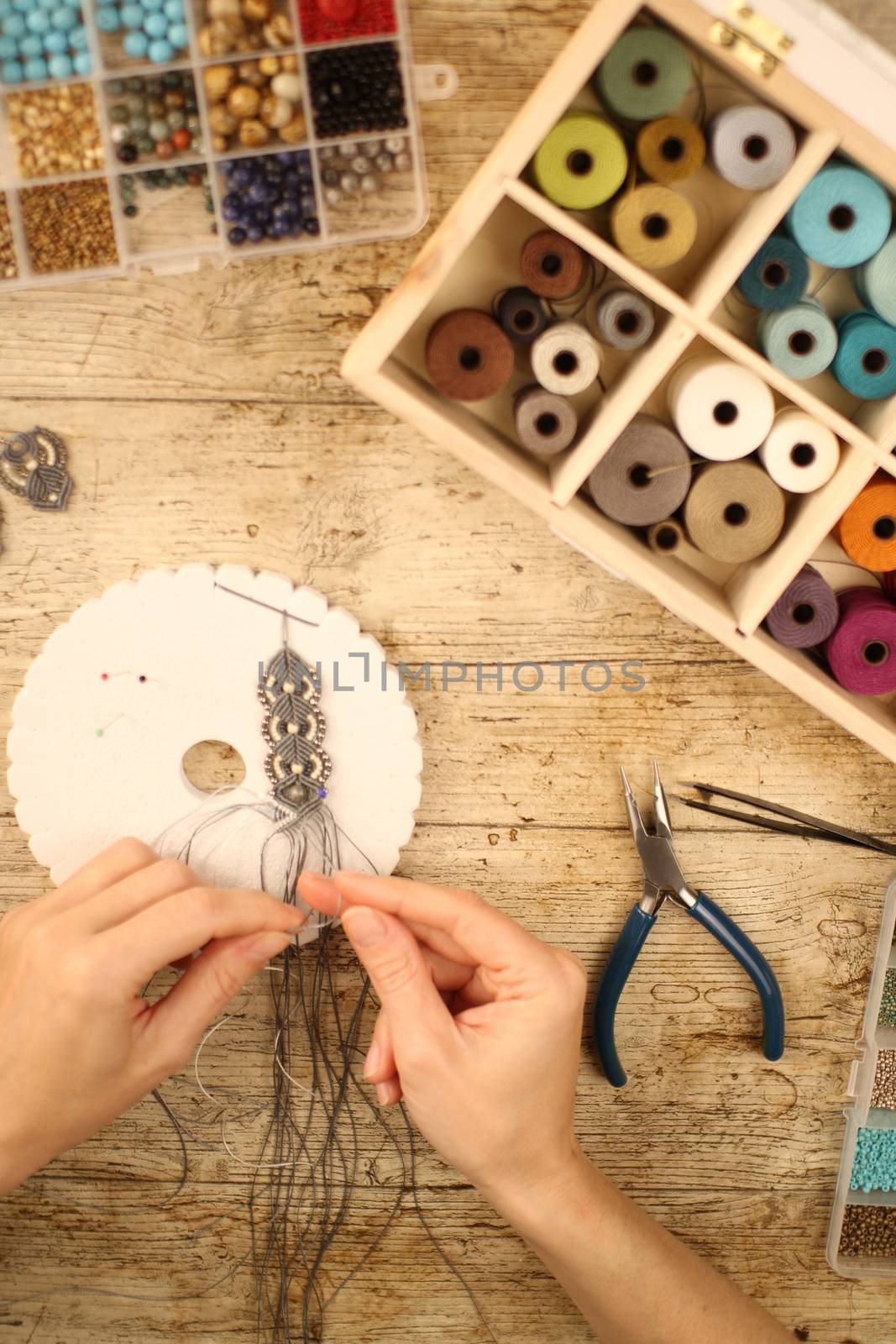 Top view of female hands making a macramé bracelet with kumihimo on a wooden table with tools, spools of thread, natural stones and colored beads by robbyfontanesi