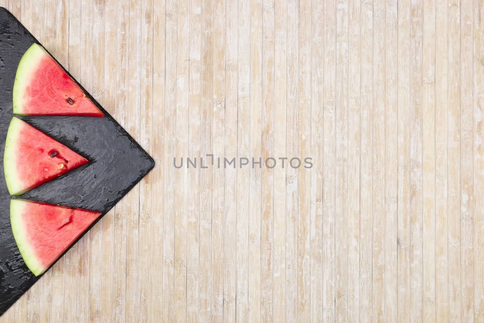 Triangular slices of watermelon on a black slate plate forming geometric games for copy space on a light wooden background by robbyfontanesi