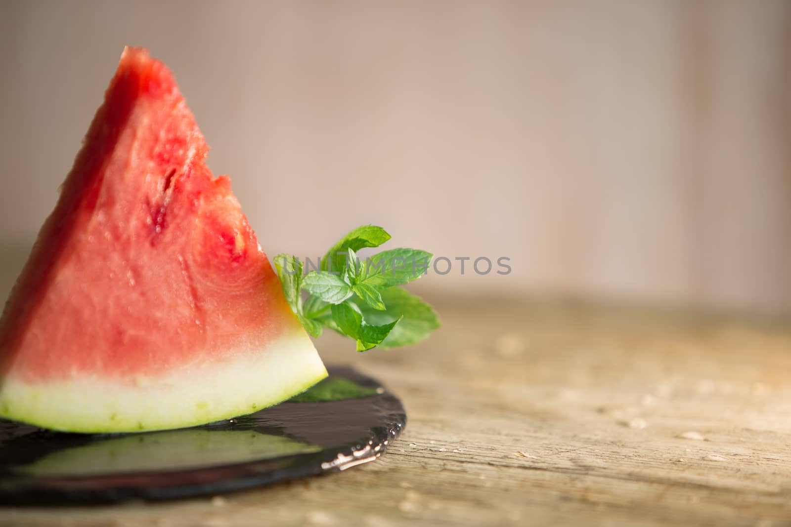 Triangular slice of watermelon standing on black plate of wet slate with sprig of fresh green mint in selective focus for copy space by robbyfontanesi