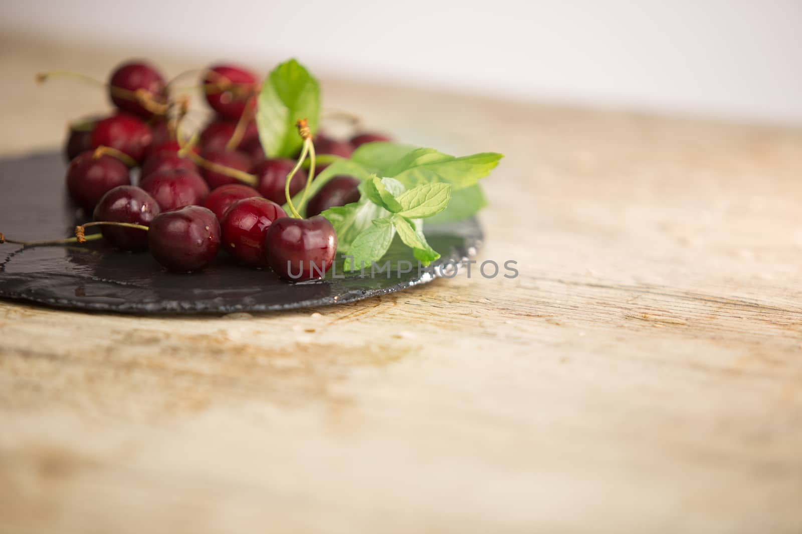 Fresh cherries on a black plate of wet slate with sprig of fresh green mint on light wooden table in selective focus for copy space by robbyfontanesi