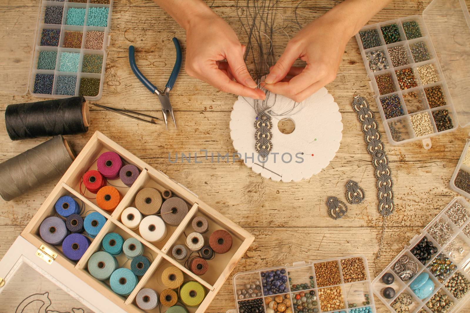 Top view of female hands making a macramé bracelet with kumihimo on a wooden table with tools, spools of thread, natural stones and colored beads