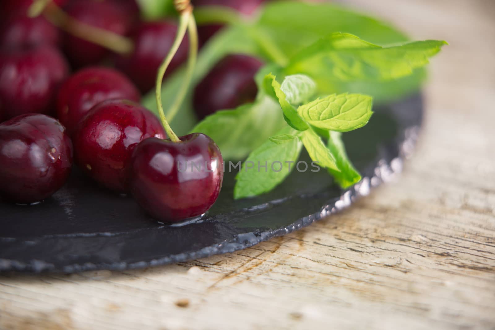 Fresh cherries on a black plate of wet slate with sprig of fresh green mint on light wooden table in selective focus for copy space by robbyfontanesi