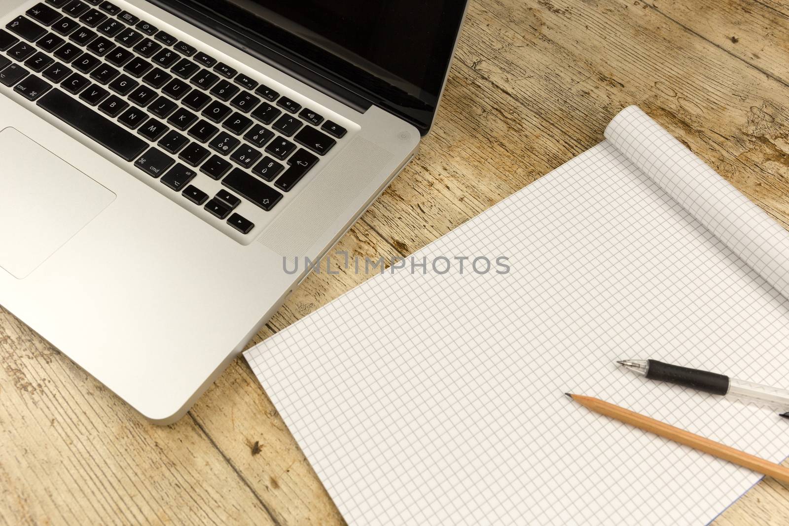 Contrast between old and new office work: shot from the top of an aluminum laptop on a wooden desk with an open checkered notebook, a pencil and a pen by robbyfontanesi