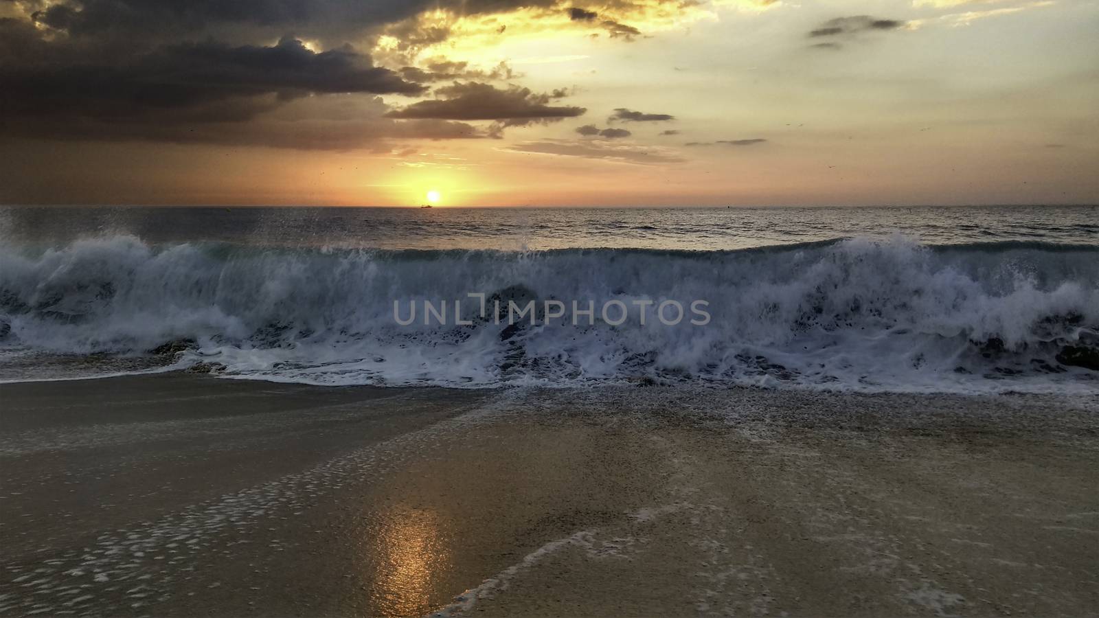 Sunset over the ocean from the shore of Nazaré beach, Playa do Norte, Portugal. The waves break on the shore reflecting the colors of the sky on the wet sand by robbyfontanesi