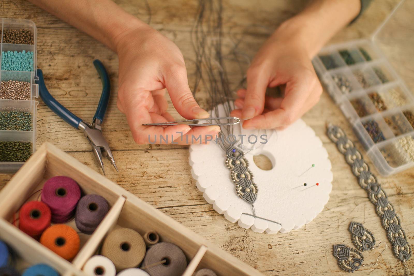 Close up of female hands making a macramé bracelet with kumihimo on a wooden table with tools, spools of thread, natural stones and colored beads