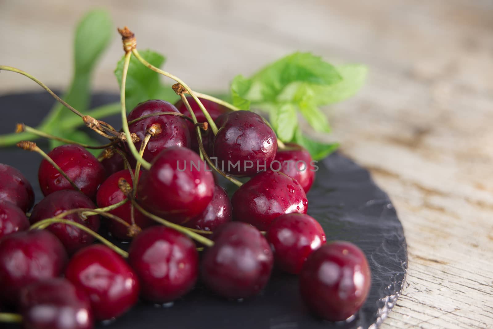 Fresh cherries on a black plate of wet slate with sprig of fresh green mint on light wooden table in selective focus for copy space by robbyfontanesi