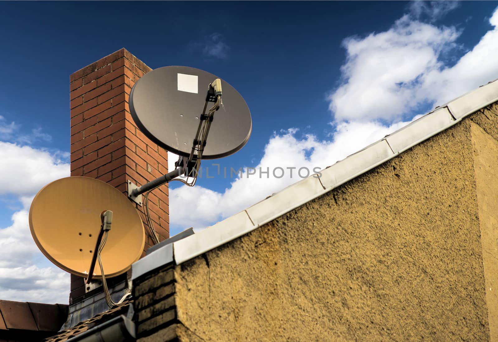 Two satellite dishes in front of a red brick chimney on the sloping roof of a house by geogif