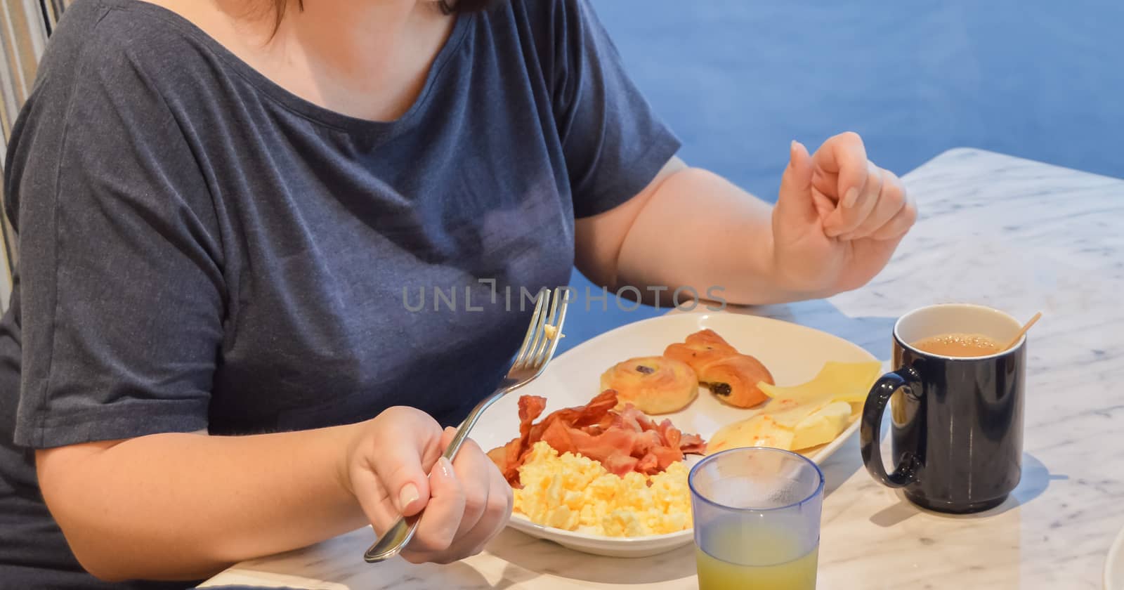 One Caucasian woman is having Breakfast in a hotel or on a cruise ship, on a plate of omelet with bacon, butter, cheese, rolls, holding a fork in her hand.