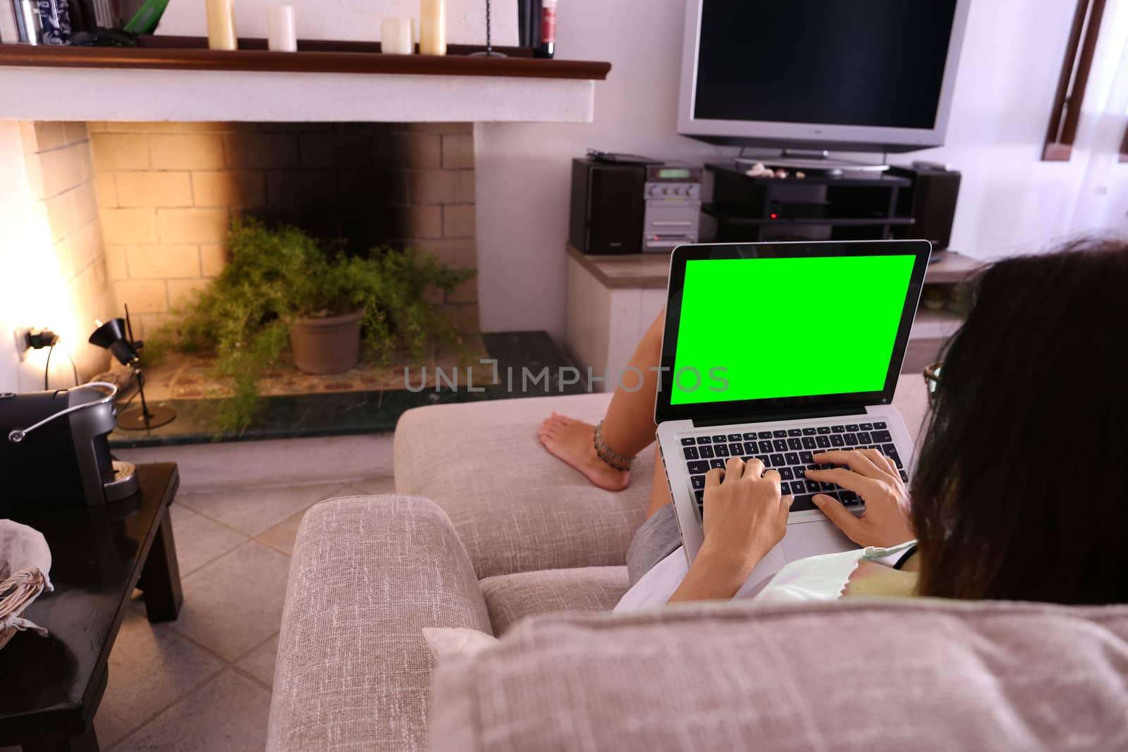 A young woman works on her laptop with the green screen at home sitting on the sofa in her living room by robbyfontanesi