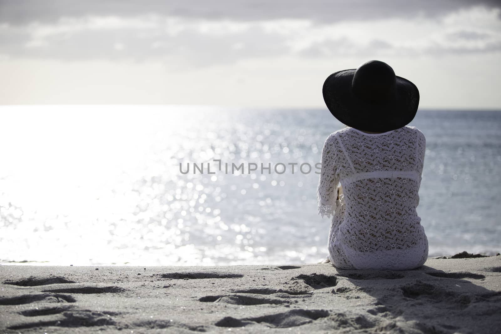 Young woman with long hair from behind sitting by the sea looks at the horizon at dawn in the wind, dressed in a white lace dress, white underwear and large black hat