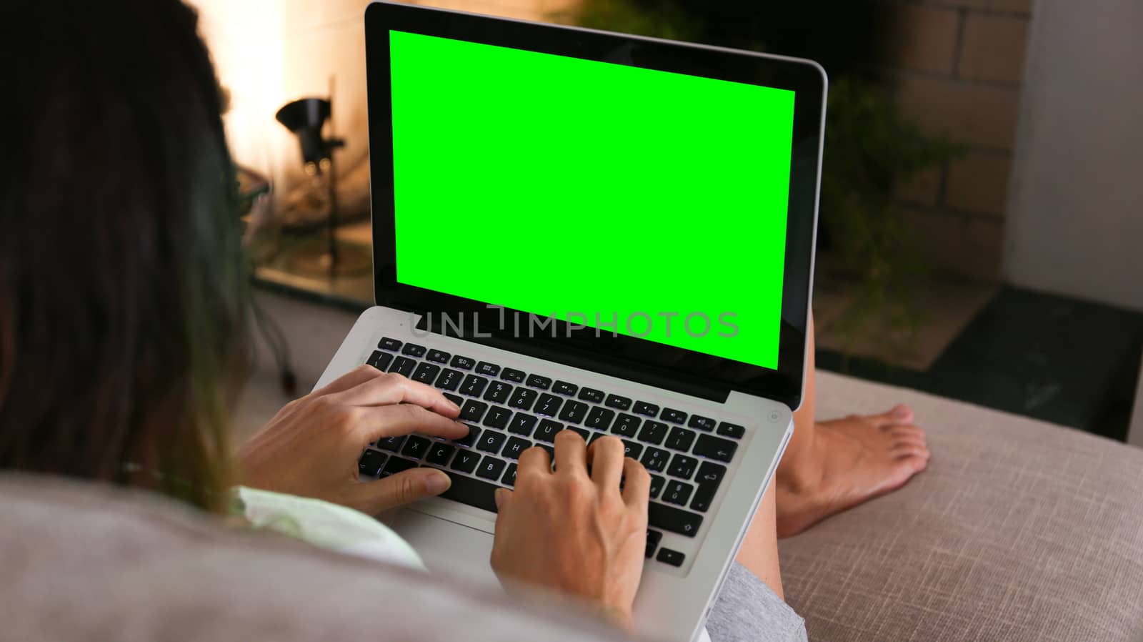 A young woman works on her laptop with the green screen at home sitting on the sofa in her living room