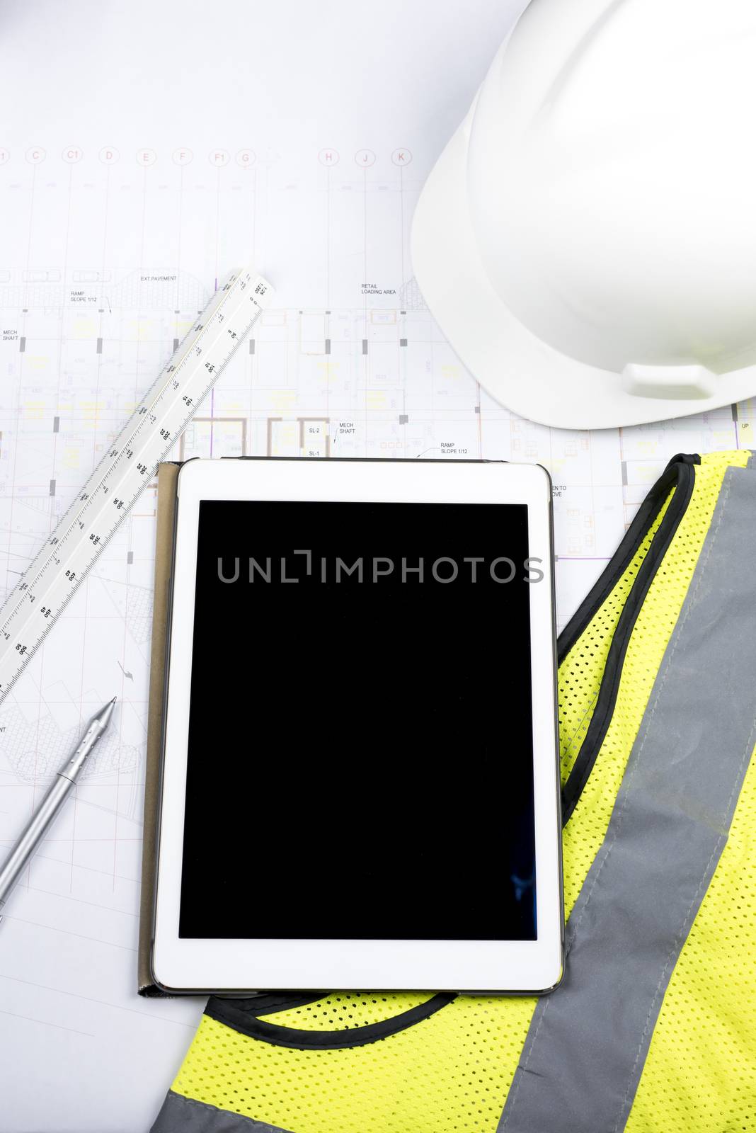 Digital tablet with black screen, hard copy of drawing, triangular scale ruler and work Personal Protective equipment (safety yellow fluorescent gilet and white helmet)