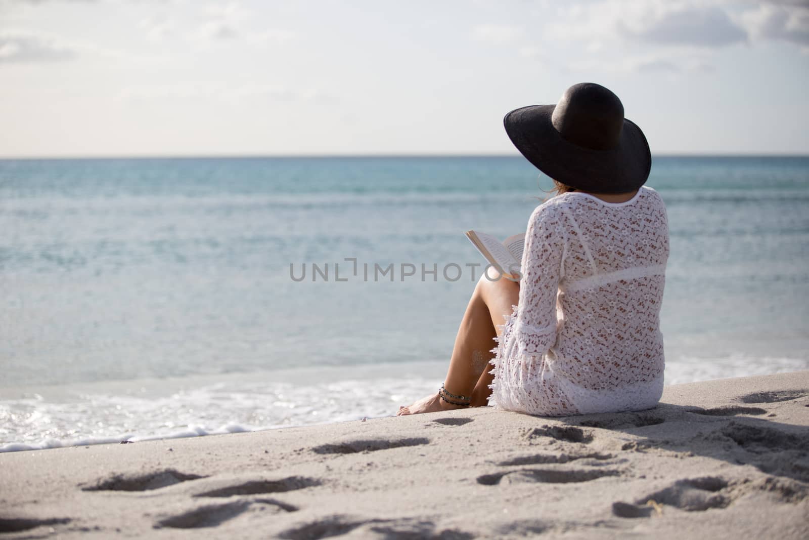 Young woman with long hair from behind sitting by the sea reads a book at dawn in the wind, dressed in a white lace dress, white underwear and large black hat by robbyfontanesi