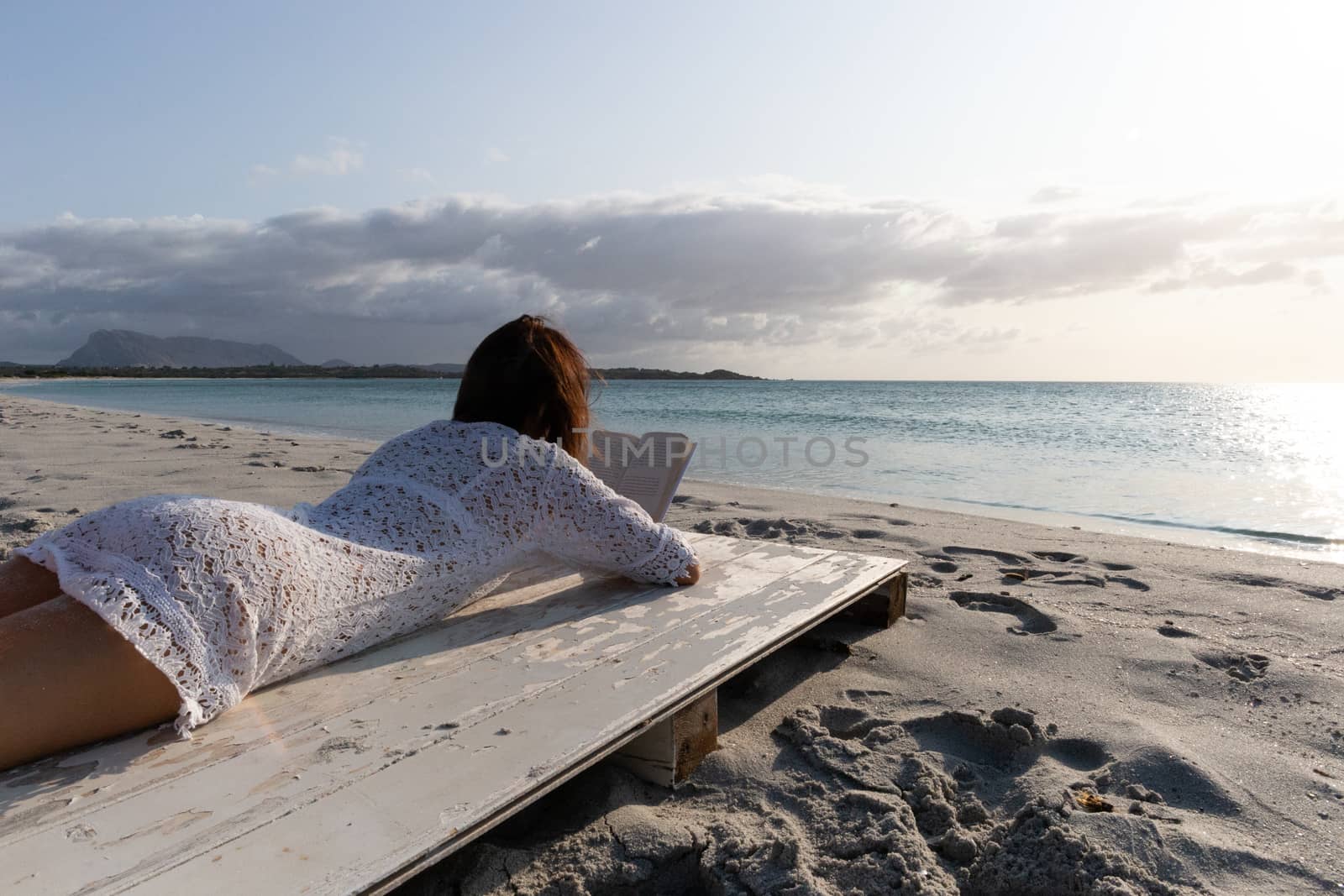 The young woman from behind lying by the sea looks at the horizon at dawn in the wind, dressed in a white lace dress and white underwear and long hair by robbyfontanesi