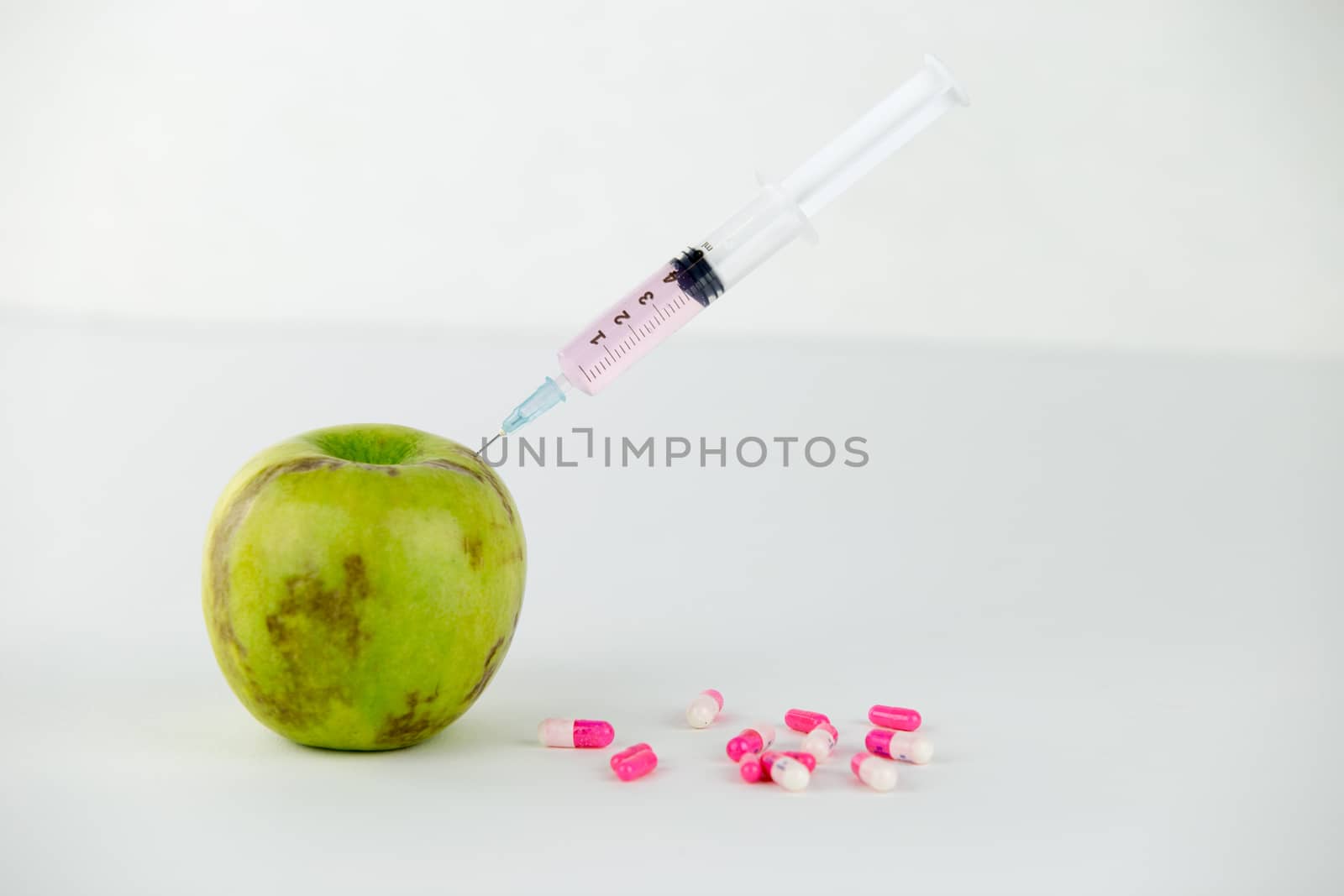 Concept: human GMO manipulation of nature and relative poisoned fruits. Close-up of an apple contaminated with a syringe threaded in and medicines on a white background