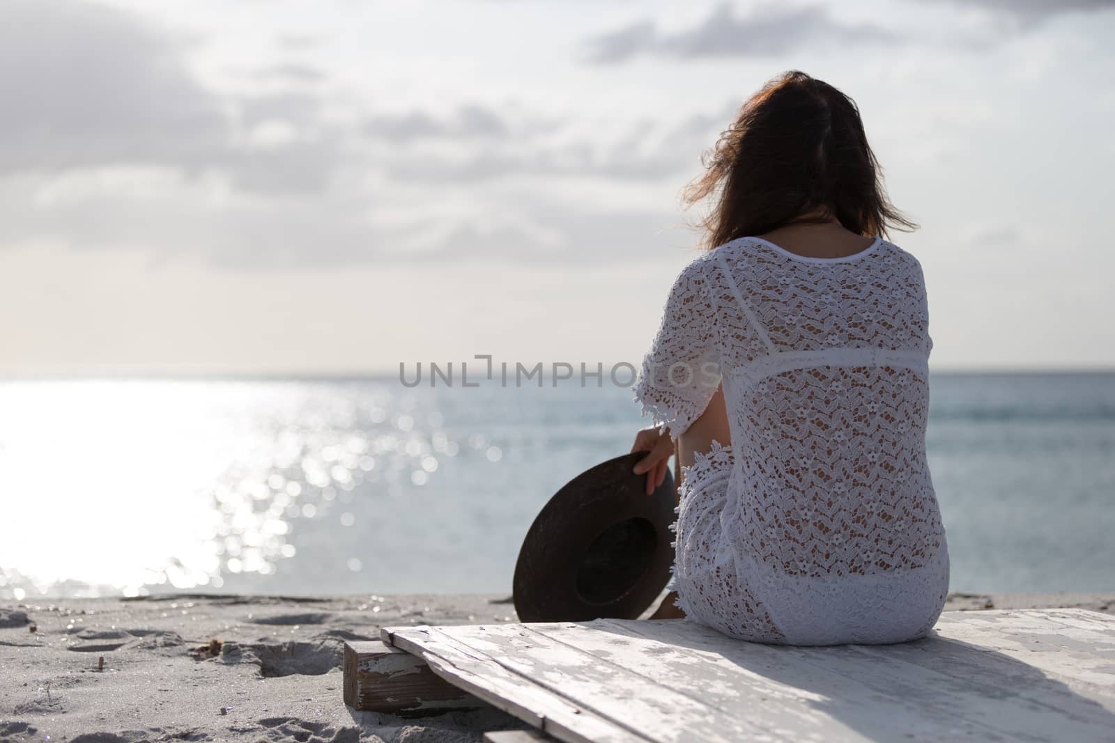 Young woman from behind sitting by the sea looks at the horizon at dawn in the wind, dressed in a white lace dress and white underwear and long hair by robbyfontanesi