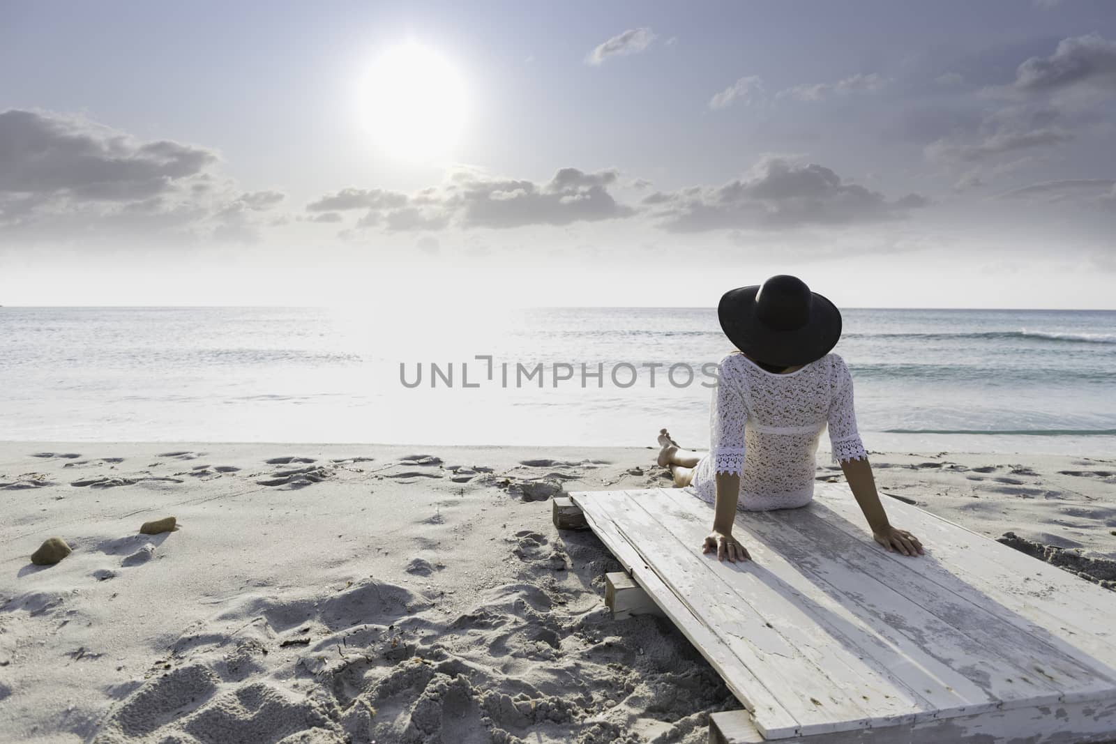 Young woman with long hair from behind sitting by the sea looks at the horizon at dawn in the wind, dressed in a white lace dress, white underwear and large black hat